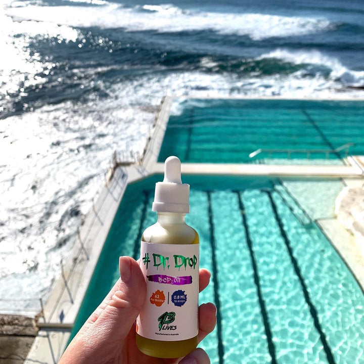 Hand holding a bottle of 13Lives Dr Drop BCP + Myrcene Oil near a pool overlooking the ocean under sunlight